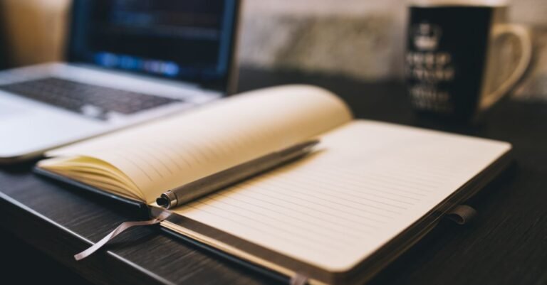 Carnet ouvert et stylo posés sur un bureau en bois à côté d’un ordinateur portable